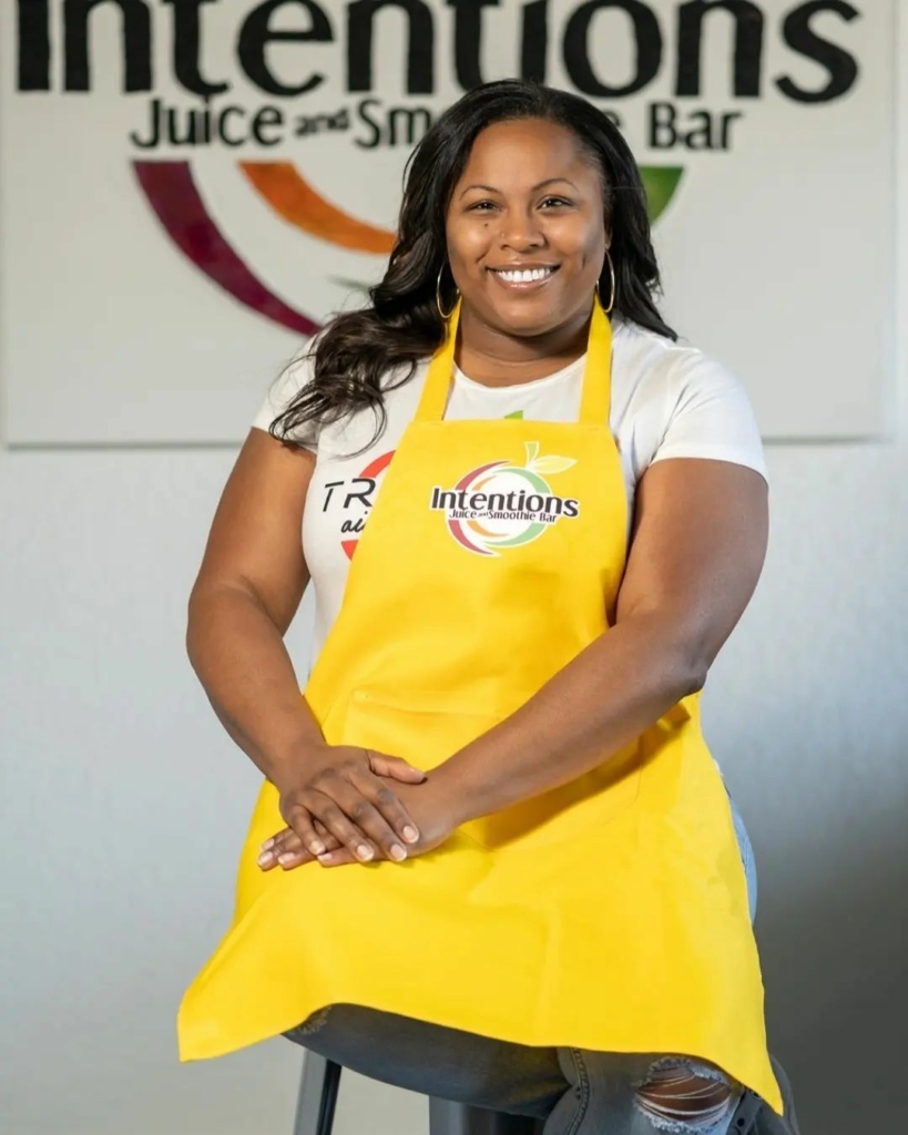 Intentions Juice Bar owner Marquita Evans, wearing a yellow apron and sitting on a stool posing for the camera with the Intentions logo on the wall behind her