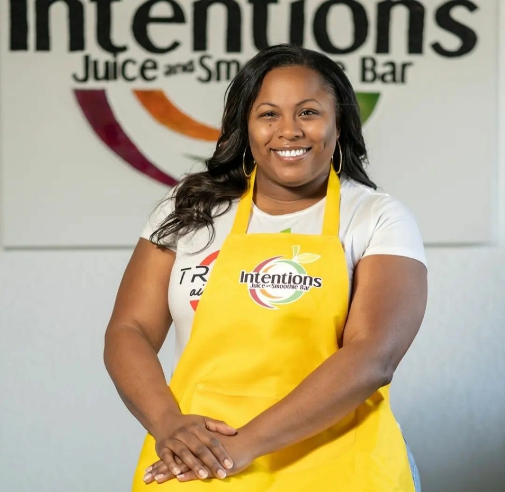 Intentions Juice Bar owner Marquita Evans, wearing a yellow apron and sitting on a stool posing for the camera with the Intentions logo on the wall behind her