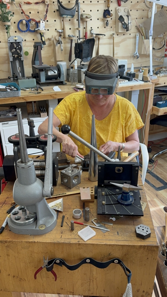 Sally works on a custom piece of jewelry in her studio. She is wearing a bright yellow shirt and magnifying glasses as she hammers a piece of metal.